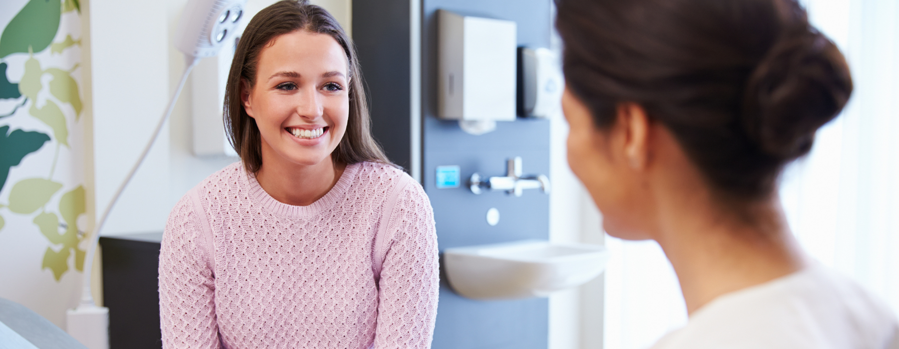 Médica sorridente em consultório, sentada frente a paciente gesticulando durante conversa empática sobre saúde.