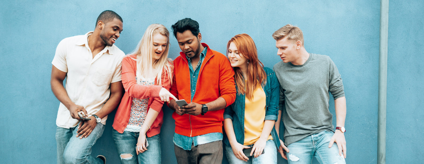 Grupo diverso de jovens encostados em uma parede azul, olhando juntos para um celular e interagindo com conteúdo digital, representando hábitos de comunicação e compartilhamento da geração Z.